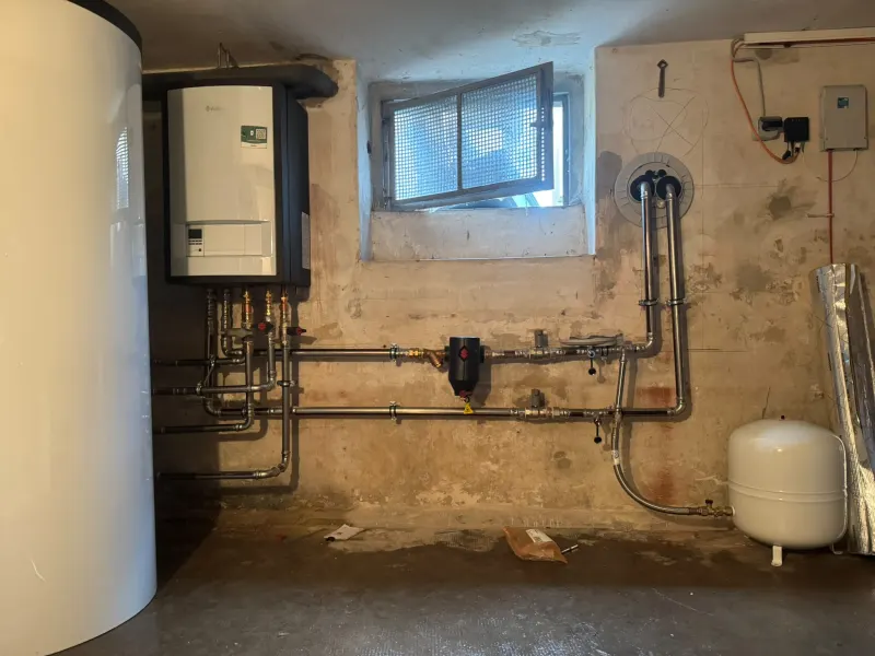 Wandgerät mit Edelstahl-Rohrleitungen, Abscheider und Ausdehnungsgefäß; die Leitungen führen zu einer Wanddurchführung unter dem Kellerfenster