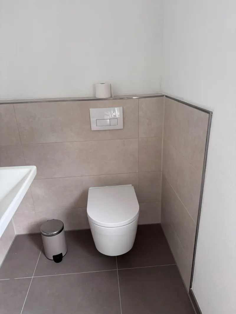 Wand-WC mit Drückerplatte vor einer halbhoch gefliesten Vorwand mit Edelstahl-Abschlussprofil